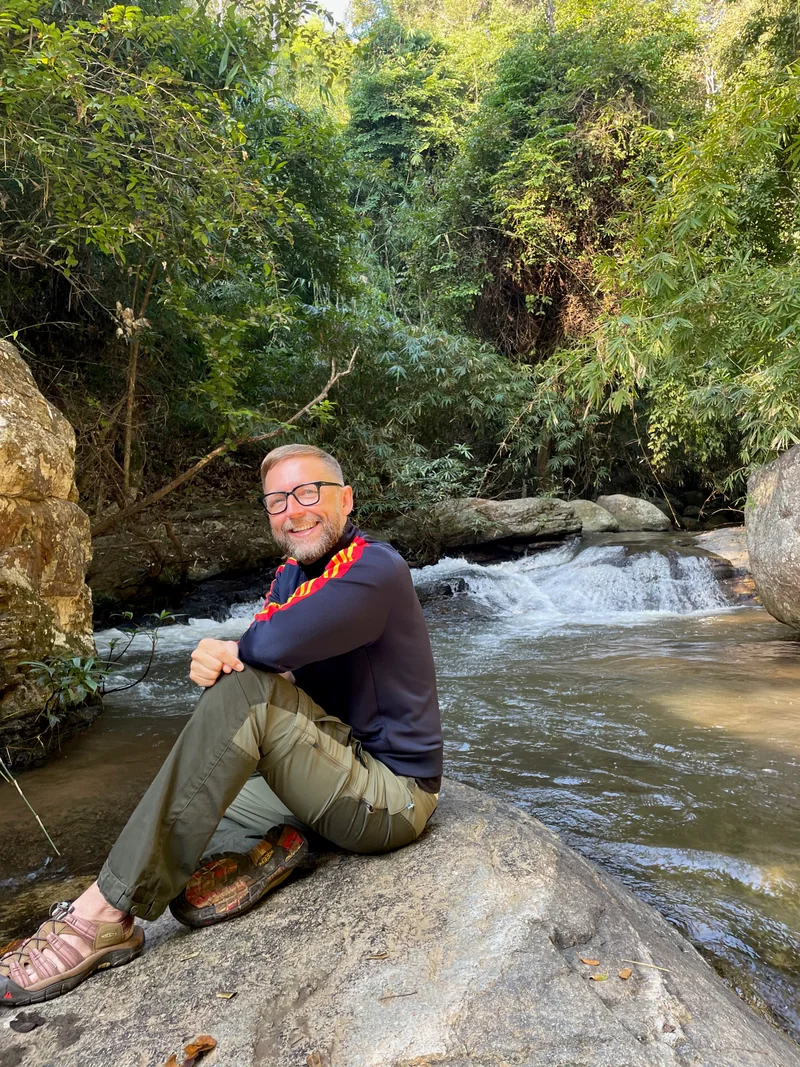 Markus Asano lacht, auf einem Felsen an einem Fluss im Grünen sitzend.